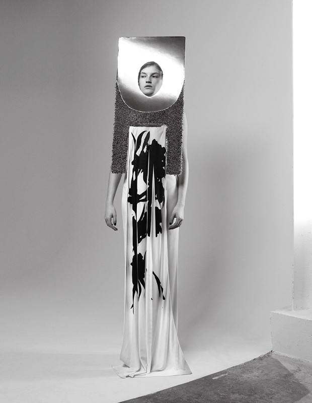 Fashion shot in black and white of woman in white gown with graphic print, head framed in reflective silver structure.