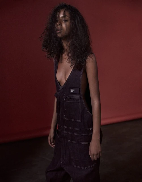 A woman with dark curly hair poses indoors wearing a sleeveless dark denim jumpsuit against a reddish-brown backdrop in a fashion editorial.