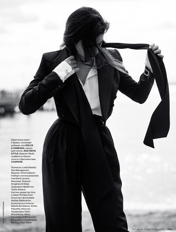 Monochrome fashion shot of a woman in a pantsuit with a scarf-tie. She is posing outdoors in front of a blurry waterscape.