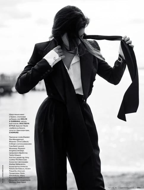 Monochrome fashion shot of a woman in a pantsuit with a scarf-tie. She is posing outdoors in front of a blurry waterscape.