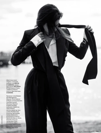 Monochrome fashion shot of a woman in a pantsuit with a scarf-tie. She is posing outdoors in front of a blurry waterscape.