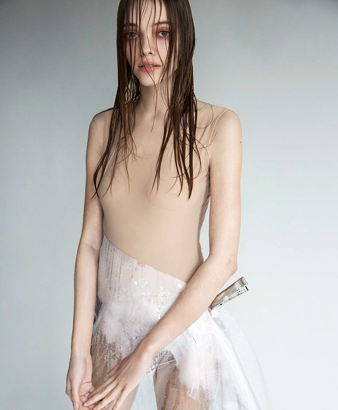 Striking fashion portrait of a young woman with wet hair and a translucent garment, set against a soft grey background, capturing minimalist style.
