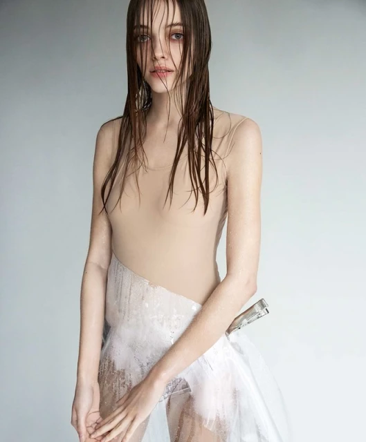 Striking fashion portrait of a young woman with wet hair and a translucent garment, set against a soft grey background, capturing minimalist style.