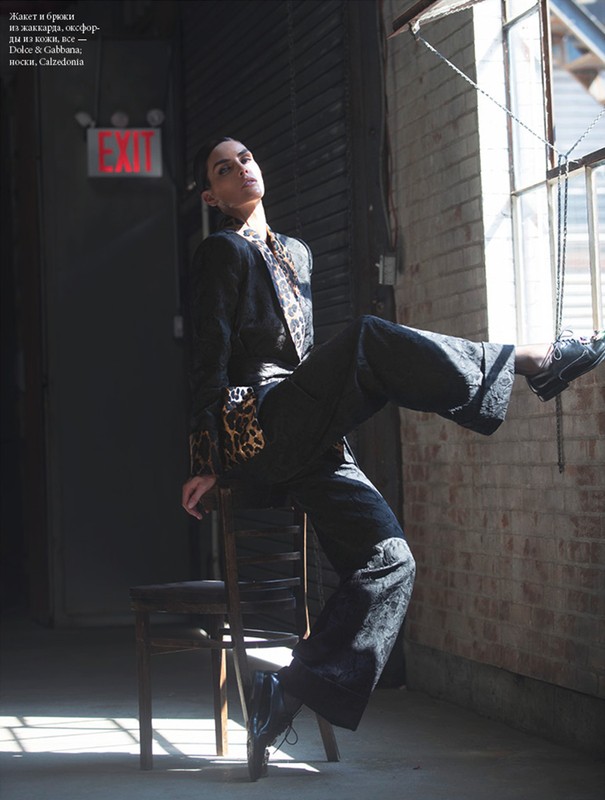 Fashionable woman in patterned blazer and wide-leg pants poses on chair in a gritty urban space near a large window.
