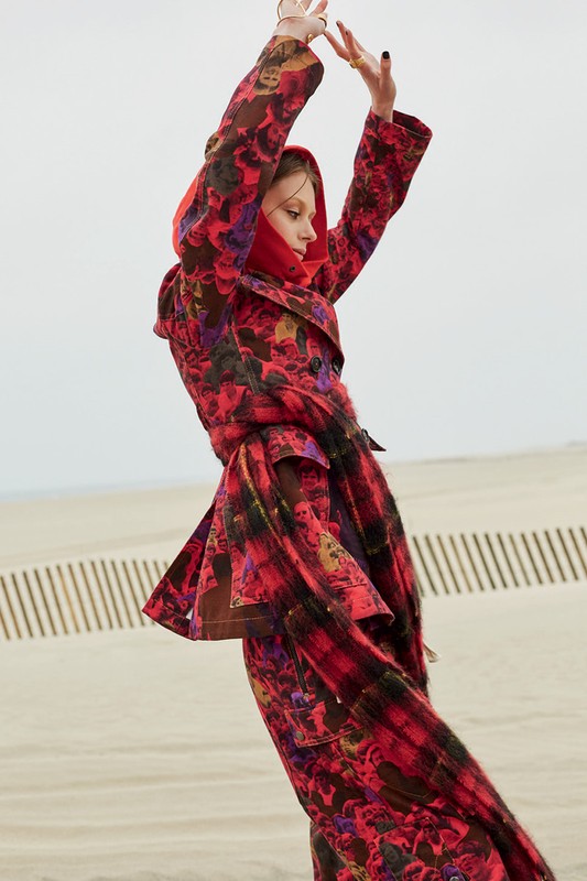 A woman in a red floral coat and matching scarf poses in a sand dune landscape, showcasing fashion and editorial style.