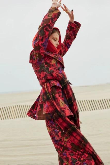 A woman in a red floral coat and matching scarf poses in a sand dune landscape, showcasing fashion and editorial style.