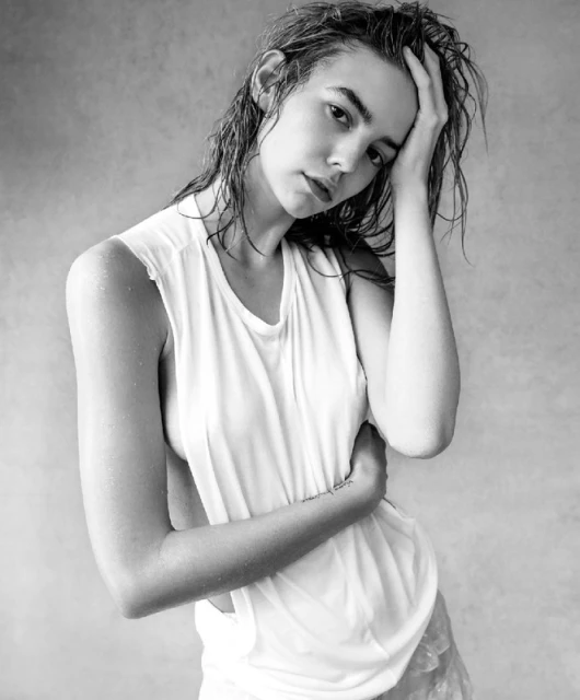 Black and white portrait of a woman with wet hair in a white sleeveless shirt, posing with a hand on her head in a studio setting.