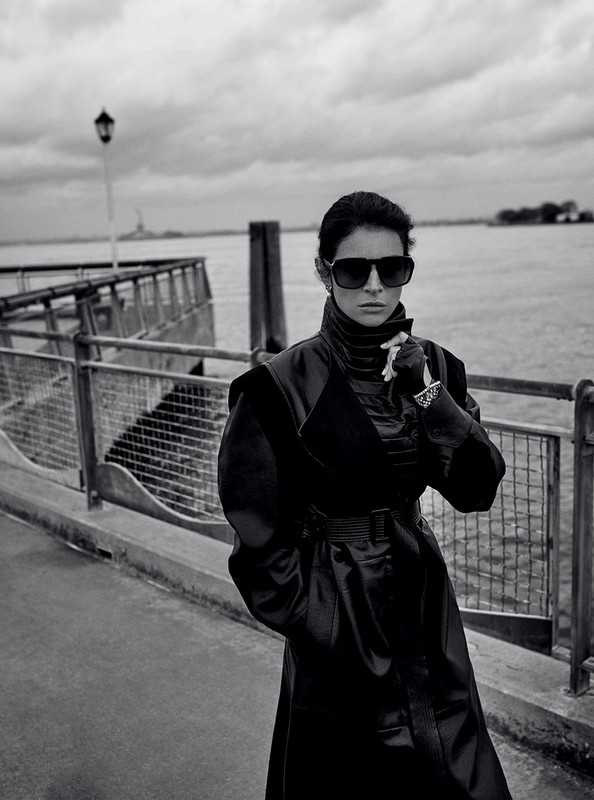 Monochrome portrait of a woman in a long dark coat and sunglasses standing by a waterfront pier
