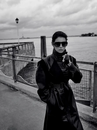 Monochrome portrait of a woman in a long dark coat and sunglasses standing by a waterfront pier