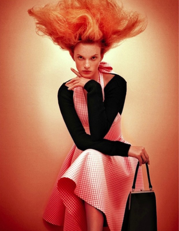 Stylish woman with vibrant red hair and a pink gingham dress poses with a classic handbag against a warm-toned backdrop.