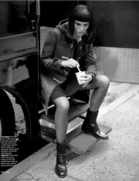 Model with dark bob hair in a wool duffle coat sitting on steps eating from a small cup, wearing black boots.