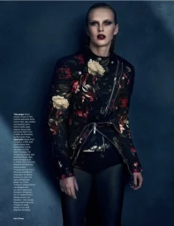 Model wearing a dark floral print biker jacket and black trousers against a textured dark background.