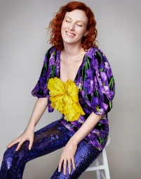 Smiling red-haired woman seated on a white stool wearing purple floral top and sequin trousers