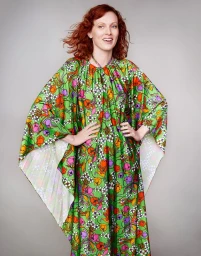 Woman with red hair wearing a vibrant green floral print caftan dress standing against a neutral grey studio background