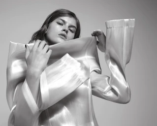 Model wearing a structured white shiny top with exaggerated sleeves against a plain grey backdrop in a black and white fashion portrait.