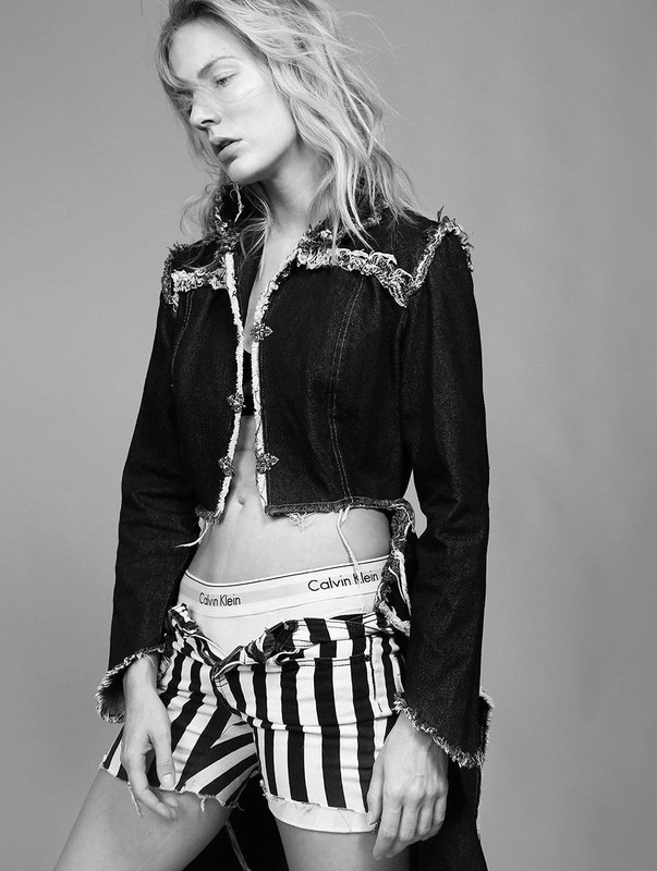 A woman poses indoors wearing a distressed denim jacket, Calvin Klein underwear, and striped shorts in a black and white fashion shot.