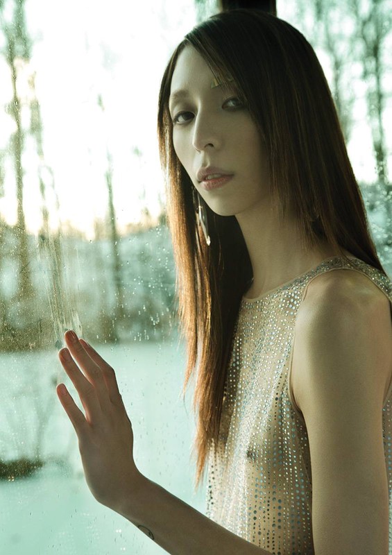 Portrait of an Asian woman in a shimmering tank top standing near a window with a snowy landscape in the background, lit by natural light.