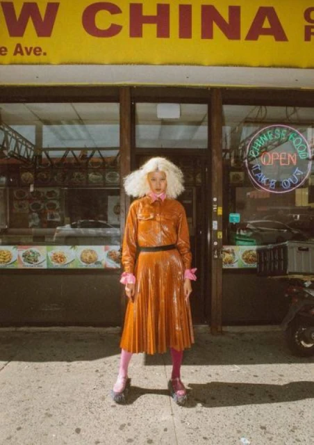A fashion-forward woman with platinum blond hair wearing a shiny orange dress and platform shoes in front of a Chinese restaurant.