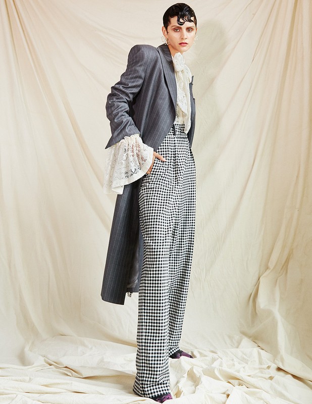 Woman in grey striped coat and black and white checkered pants stands in a studio with a cream colored backdrop, fashion shoot.