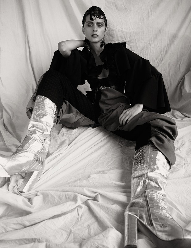 Studio shot of a young adult woman with dark hair and metallic boots, sitting in a draped studio setting. Fashion editorial in black and white.
