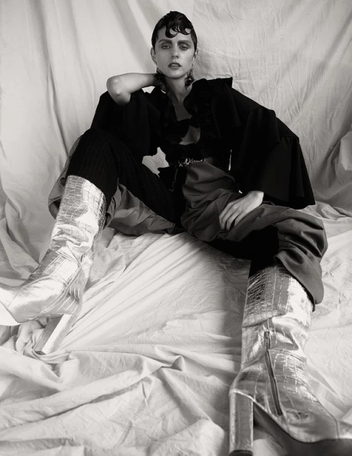 Studio shot of a young adult woman with dark hair and metallic boots, sitting in a draped studio setting. Fashion editorial in black and white.