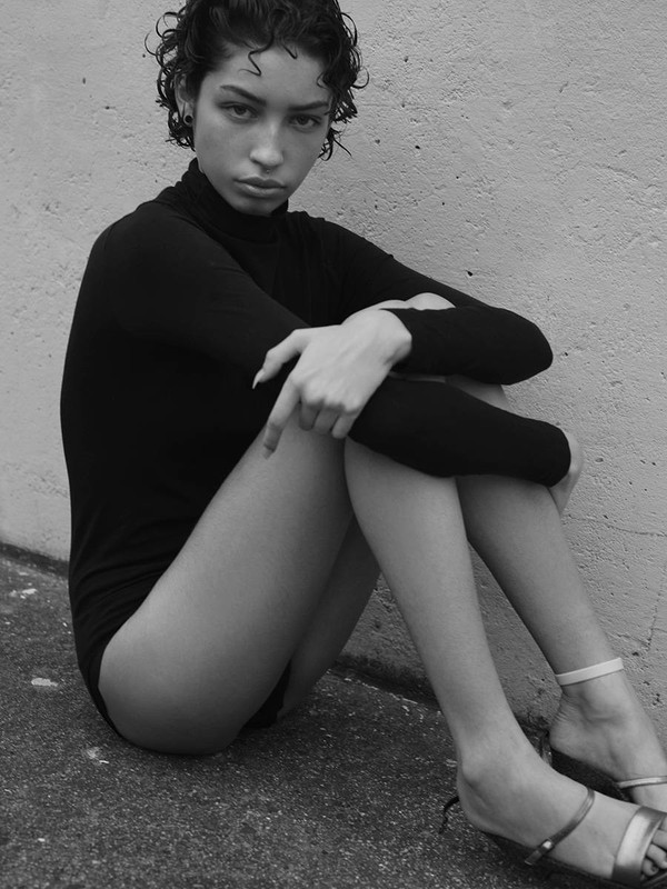 Young woman with short curly hair in a black bodysuit sitting in front of a wall looking at the camera in a studio shot.