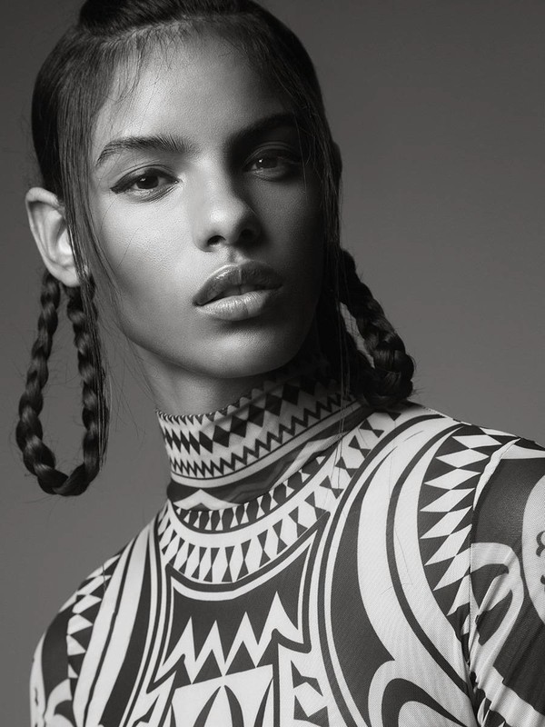 Black and white studio portrait of a woman with braided hair wearing a patterned, high-necked top against a solid gray background.