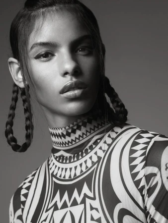 Black and white studio portrait of a woman with braided hair wearing a patterned, high-necked top against a solid gray background.
