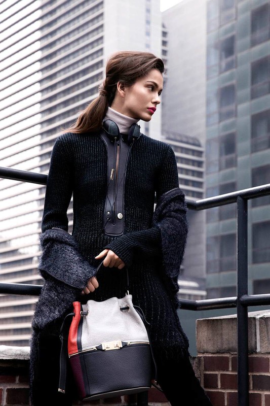 Woman in black sweater and gray bucket bag poses on building rooftop, with tall buildings in the background on overcast day.
