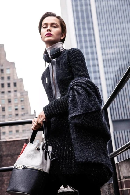Stylish woman in urban setting with a designer handbag, scarf, and headphones; city buildings in background, fashion editorial.