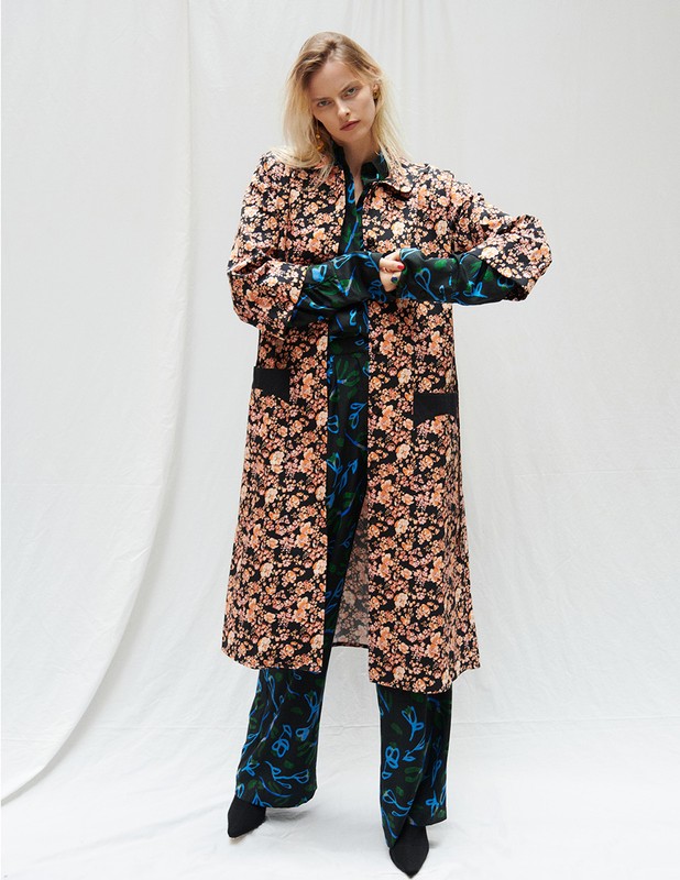 A woman in a floral print coat and pants poses in a bright studio with white draping in the background for a fashion editorial shot.