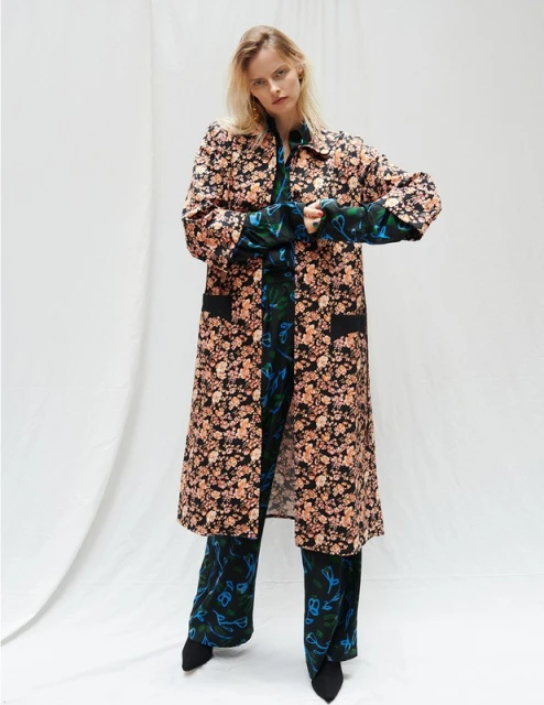 A woman in a floral print coat and pants poses in a bright studio with white draping in the background for a fashion editorial shot.