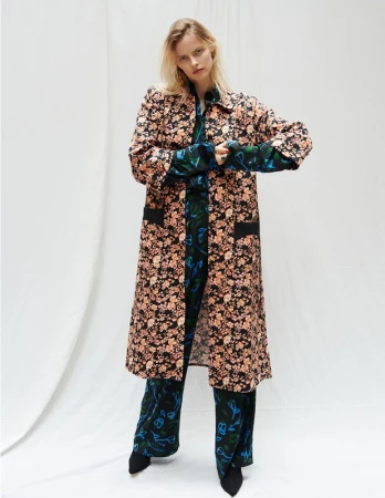 A woman in a floral print coat and pants poses in a bright studio with white draping in the background for a fashion editorial shot.