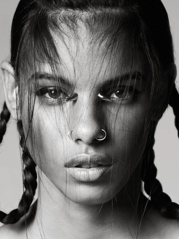 Close-up portrait of a woman with braided hair, nose rings, and metal accents under her eyes in a black and white studio shot.