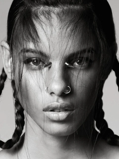 Close-up portrait of a woman with braided hair, nose rings, and metal accents under her eyes in a black and white studio shot.