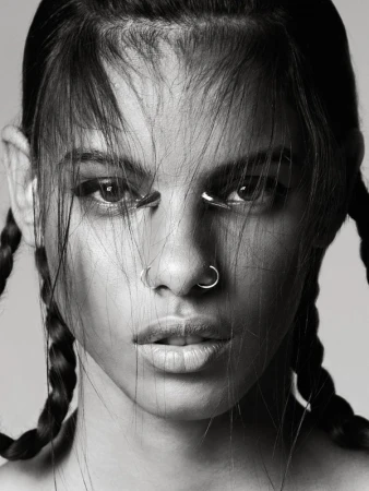 Close-up portrait of a woman with braided hair, nose rings, and metal accents under her eyes in a black and white studio shot.