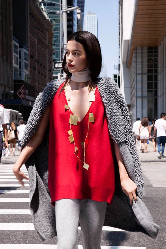 A woman in a red top and gray cardigan walks down a city street with tall buildings under a bright sky on a sunny day.
