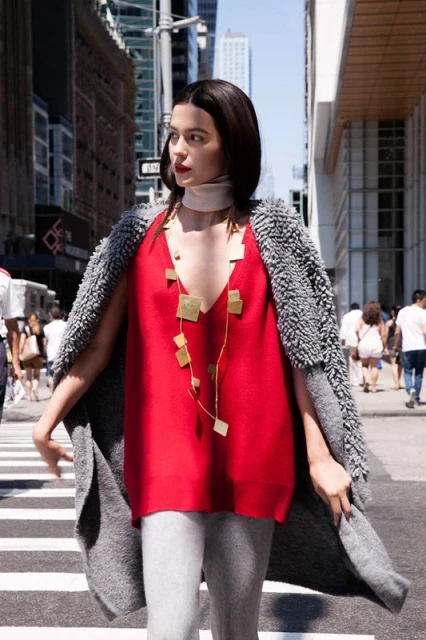 A woman in a red top and gray cardigan walks down a city street with tall buildings under a bright sky on a sunny day.