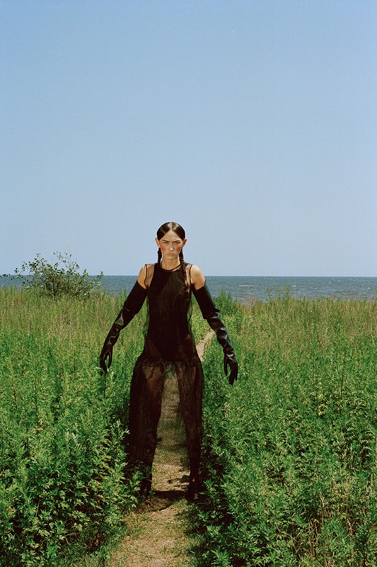 Woman in sheer black dress stands in a green field, wearing long black gloves and boots on a path leading to the ocean under a blue sky.