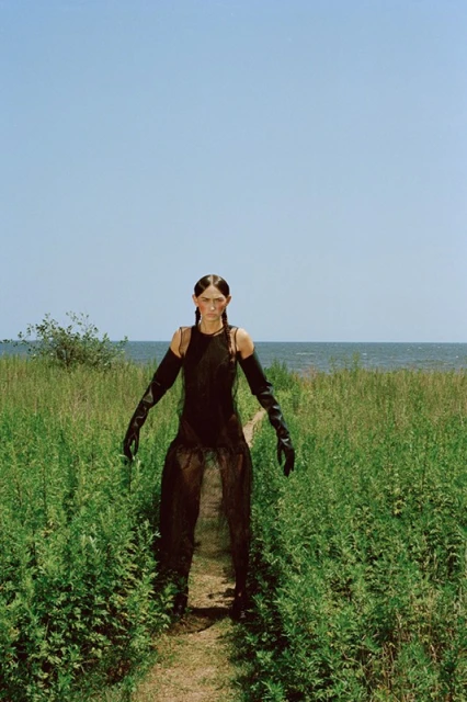 Woman in sheer black dress stands in a green field, wearing long black gloves and boots on a path leading to the ocean under a blue sky.
