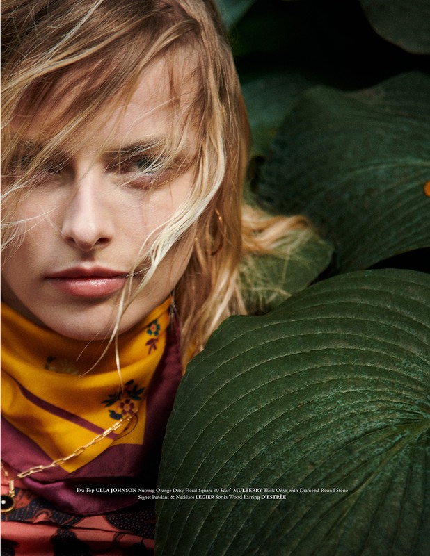 Close-up of a blonde woman with a patterned scarf, gold chain, and leafy green plants, styled in a lush and natural environment.