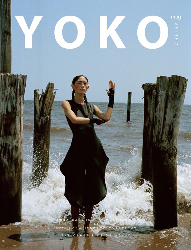 Yoko Mag Online cover featuring a slender model in a black dress standing on a sandy beach amid weathered wooden posts and crashing waves.