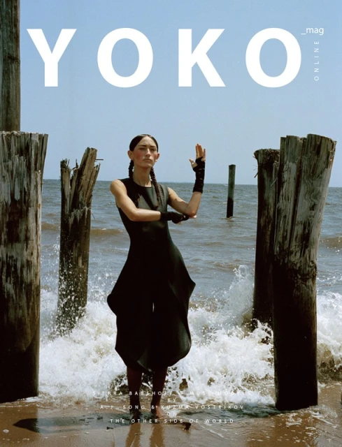 Yoko Mag Online cover featuring a slender model in a black dress standing on a sandy beach amid weathered wooden posts and crashing waves.