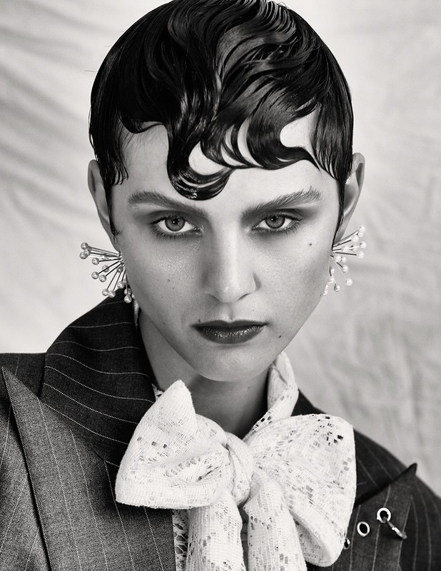 Black and white portrait of a woman with slicked-back hair wearing statement earrings, a striped blazer, and a lace bow tie.