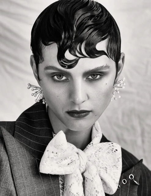 Black and white portrait of a woman with slicked-back hair wearing statement earrings, a striped blazer, and a lace bow tie.