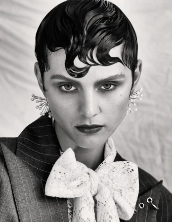Black and white portrait of a woman with slicked-back hair wearing statement earrings, a striped blazer, and a lace bow tie.