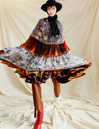 Model in layered patterned dress and black hat wearing red boots against a draped backdrop