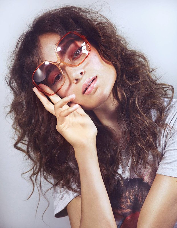 Brunette woman poses in a gray t-shirt and brown tinted, octagon-shaped sunglasses, hand near face in a studio with soft lighting.