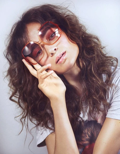 Brunette woman poses in a gray t-shirt and brown tinted, octagon-shaped sunglasses, hand near face in a studio with soft lighting.