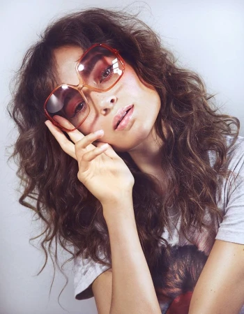Brunette woman poses in a gray t-shirt and brown tinted, octagon-shaped sunglasses, hand near face in a studio with soft lighting.